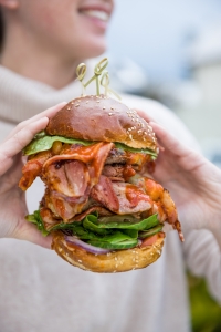 A diner at the Tradewinds Restaurant holds a large burger made with a sesame bun and loaded with bacon, beef patties, lettuce, cucumber, red onion, and spicy sauce